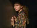 Justyna Wasilewska w kolorowej kurtce opiera się o ciemną ścianę, jej twarz oświetla cień.