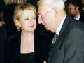 Ewa Holoubek w czarnej sukni i marynarce rozmawia z mężczyzną w garniturze.