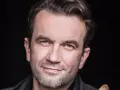 Tomasz Kot zamyślony, z lekkim uśmiechem na twarzy, na czarnym tle.