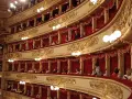 Wnętrze teatru La Scala, z widownią w lożach ozdobionych złotem i czerwonymi zasłonami.