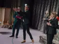 Arkadiusz Brykalski w garniturze gestykuluje na scenie, obok niego mężczyzna z teczką. W tle kobiety w eleganckich strojach.