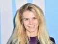 Uśmiechnięta Marta Staśmierska z długimi blond włosami, w fioletowej bluzce, na tle niebiesko-białych pasów.