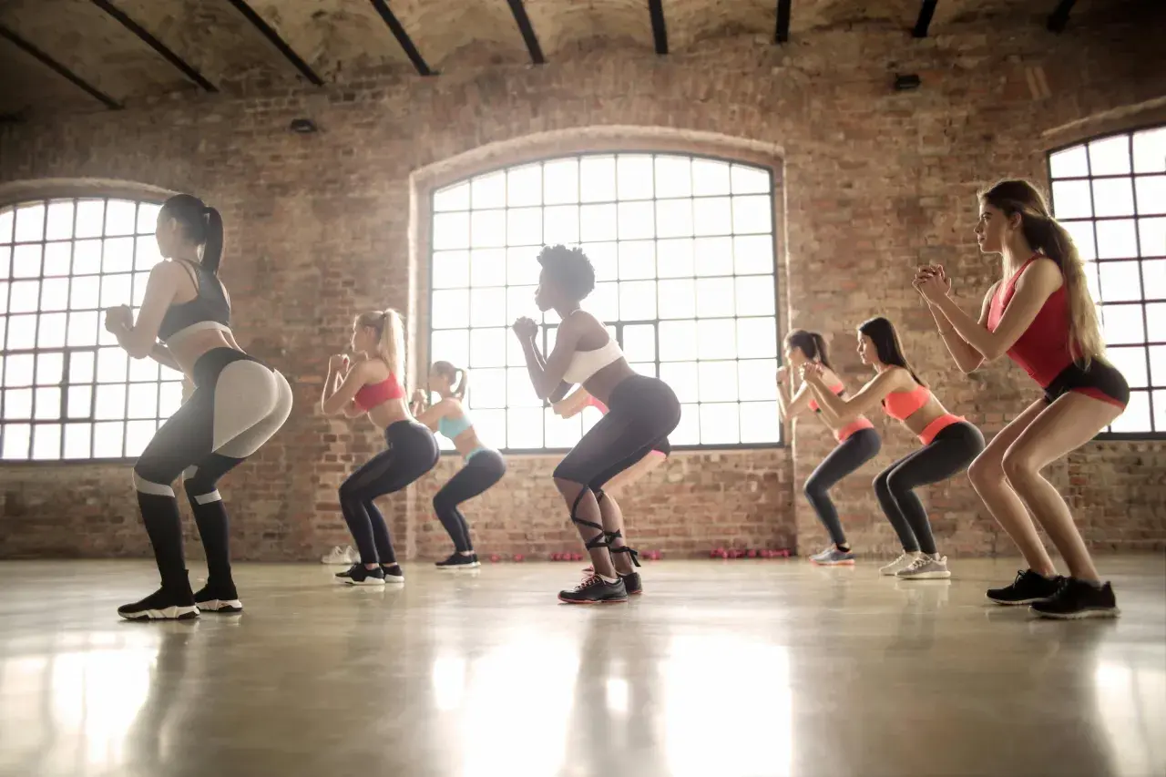 Grupa kobiet ćwiczy w rytm muzyki, wykonując przysiady. To właśnie zumba – świetna zabawa i trening w jednym!