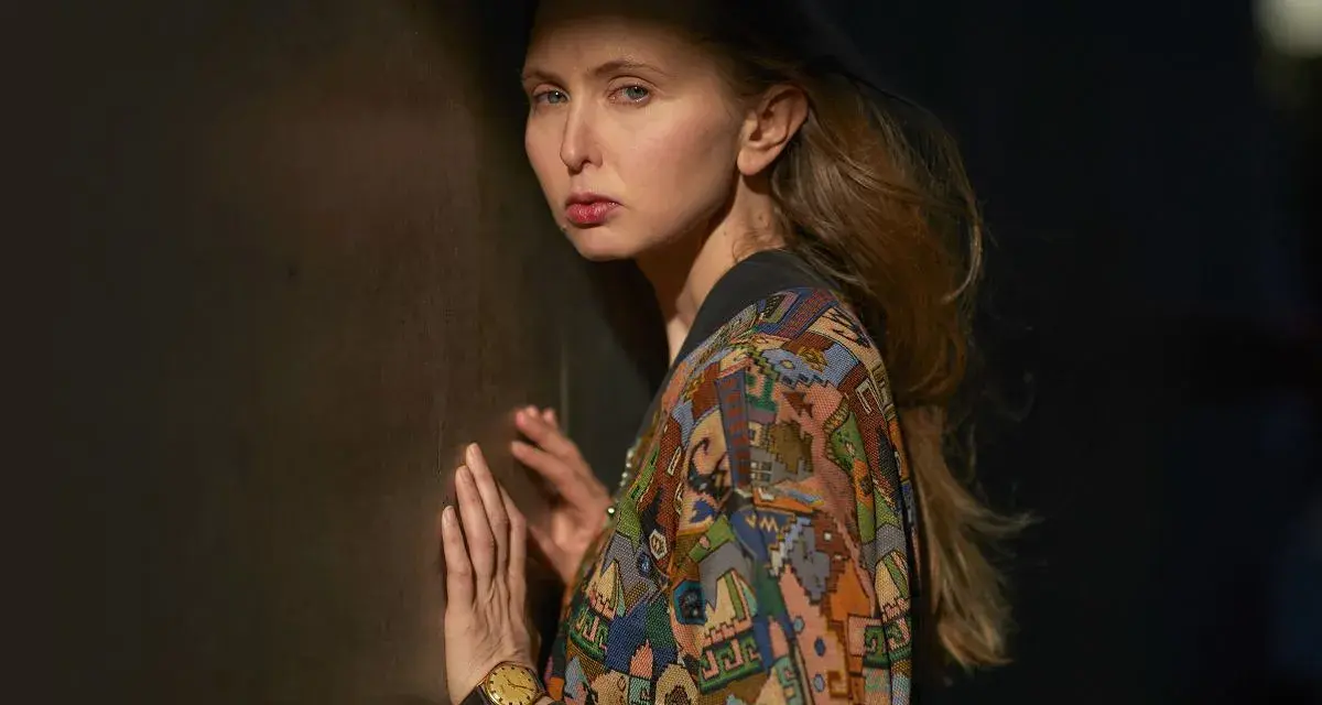 Justyna Wasilewska w kolorowej kurtce opiera się o ciemną ścianę, jej twarz oświetla cień.