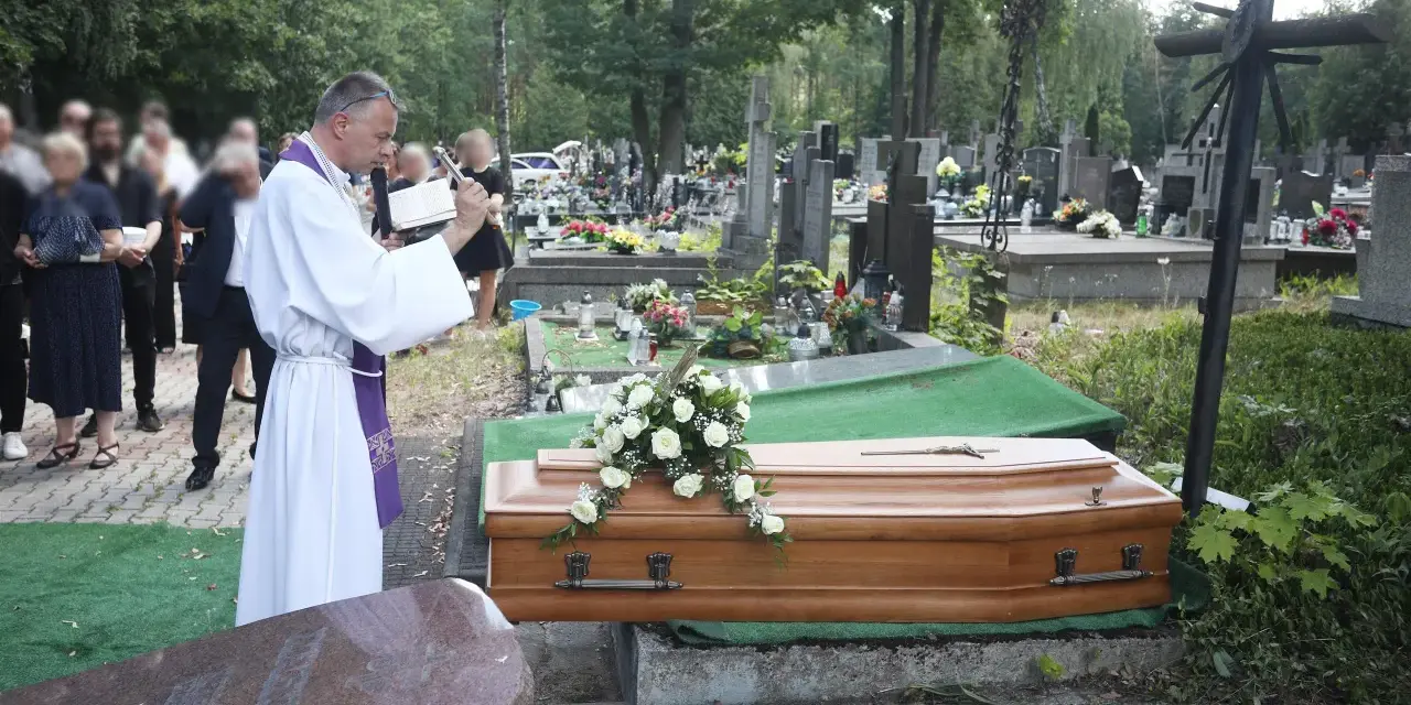 Wojciech Magnuski żegna zmarłego na cmentarzu. Ksiądz w białej szacie czyta z księgi przy trumnie ozdobionej białymi różami.