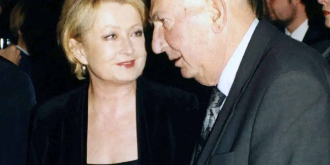 Ewa Holoubek w czarnej sukni i marynarce rozmawia z mężczyzną w garniturze.
