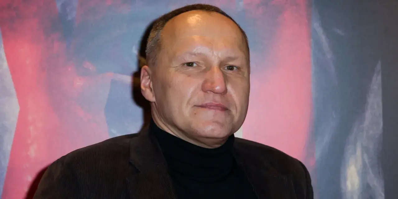 Portret Mariusza Grzegorzka w czarnym golfie i brązowej marynarce na tle abstrakcyjnego wzoru.