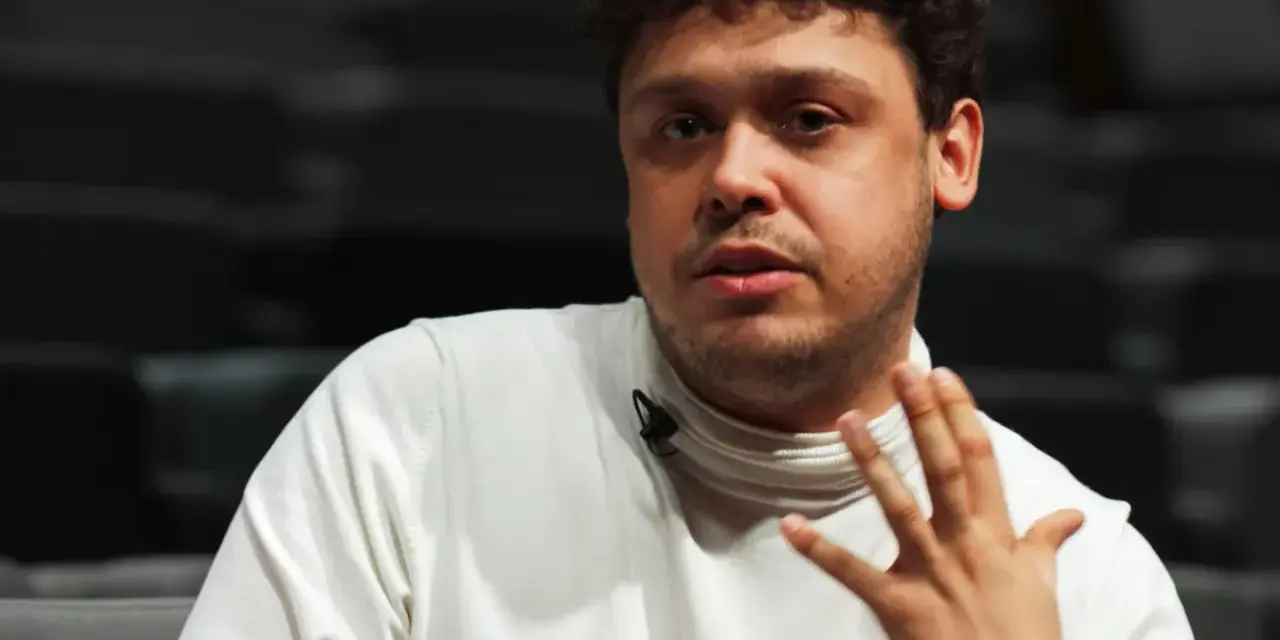 Mężczyzna z kręconymi włosami, Jakub Skrzywanek, w białym golfie gestykuluje podczas rozmowy.