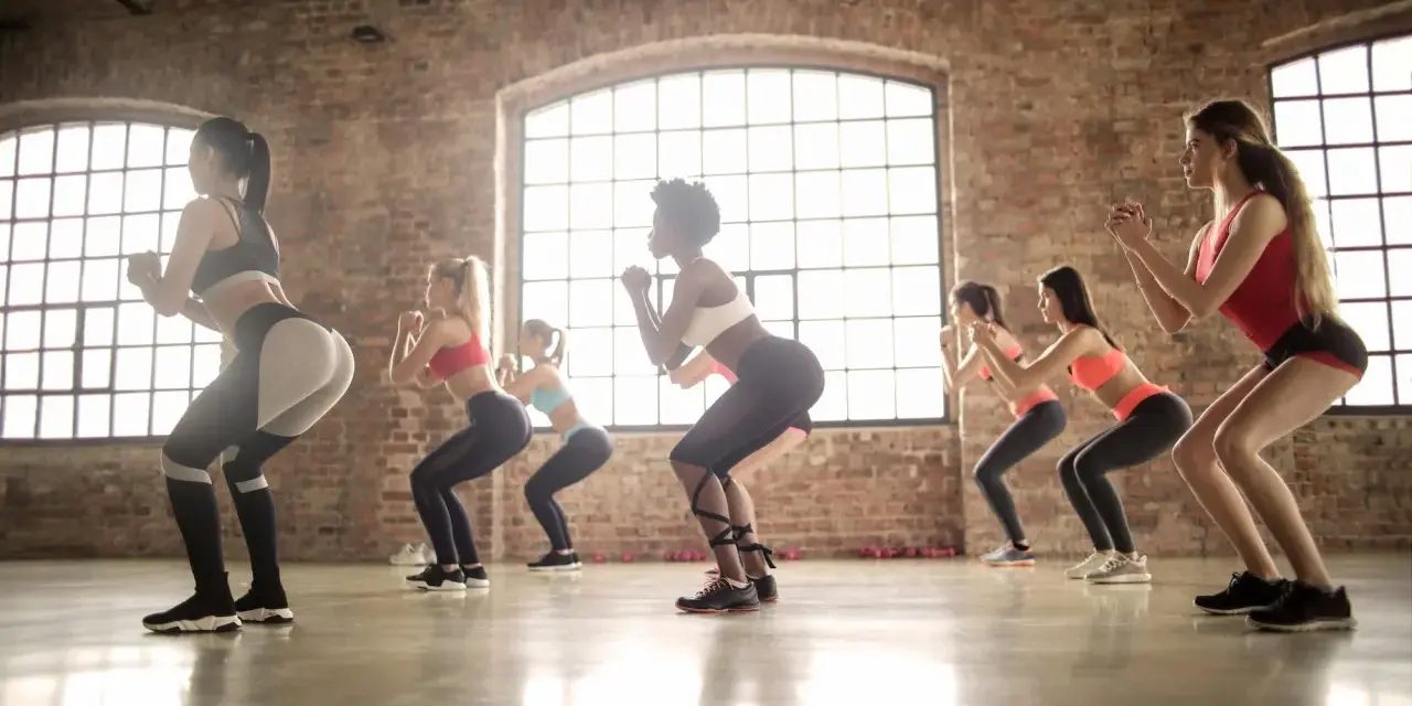Grupa kobiet ćwiczy w rytm muzyki, wykonując przysiady. To właśnie zumba – świetna zabawa i trening w jednym!