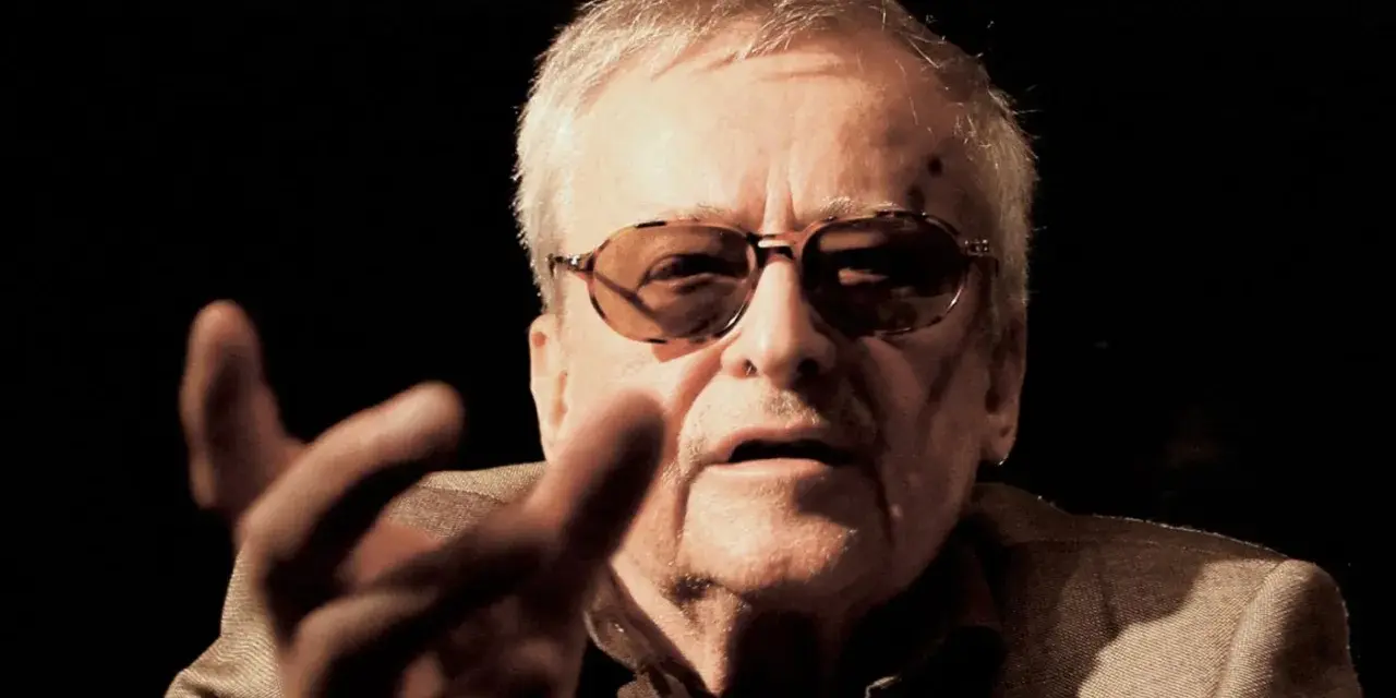 Jerzy Jarocki w okularach, z gestem dłoni, w ciemnym tle.
