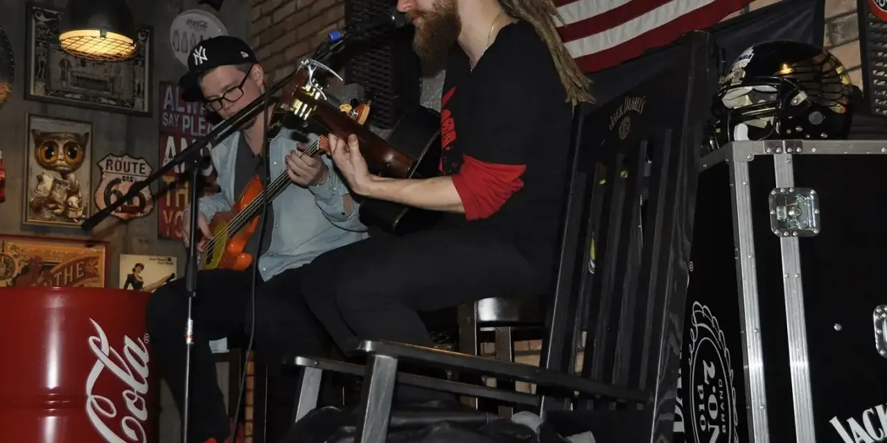 Dwóch muzyków gra na gitarach w klimatycznym wnętrzu. Jeden z nich, muzyk grający do kotleta, ma dredy i brodę.