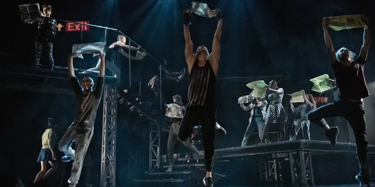 Na scenie "Metro Janusza Józefowicza" tancerze wznoszą gazety, tworząc dynamiczną choreografię.