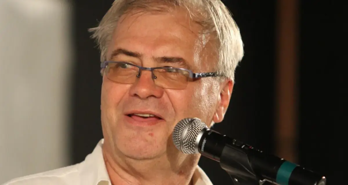 Andrzej Błaszczak, aktor, w okularach i białej koszuli, przemawia do mikrofonu.