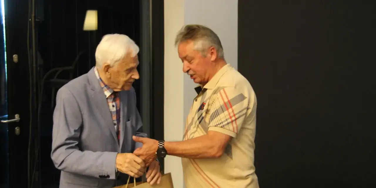Zdzisław Kuźniar syn, starszy pan w niebieskiej marynarce, wręcza prezent młodszemu mężczyźnie w beżowej koszulce.