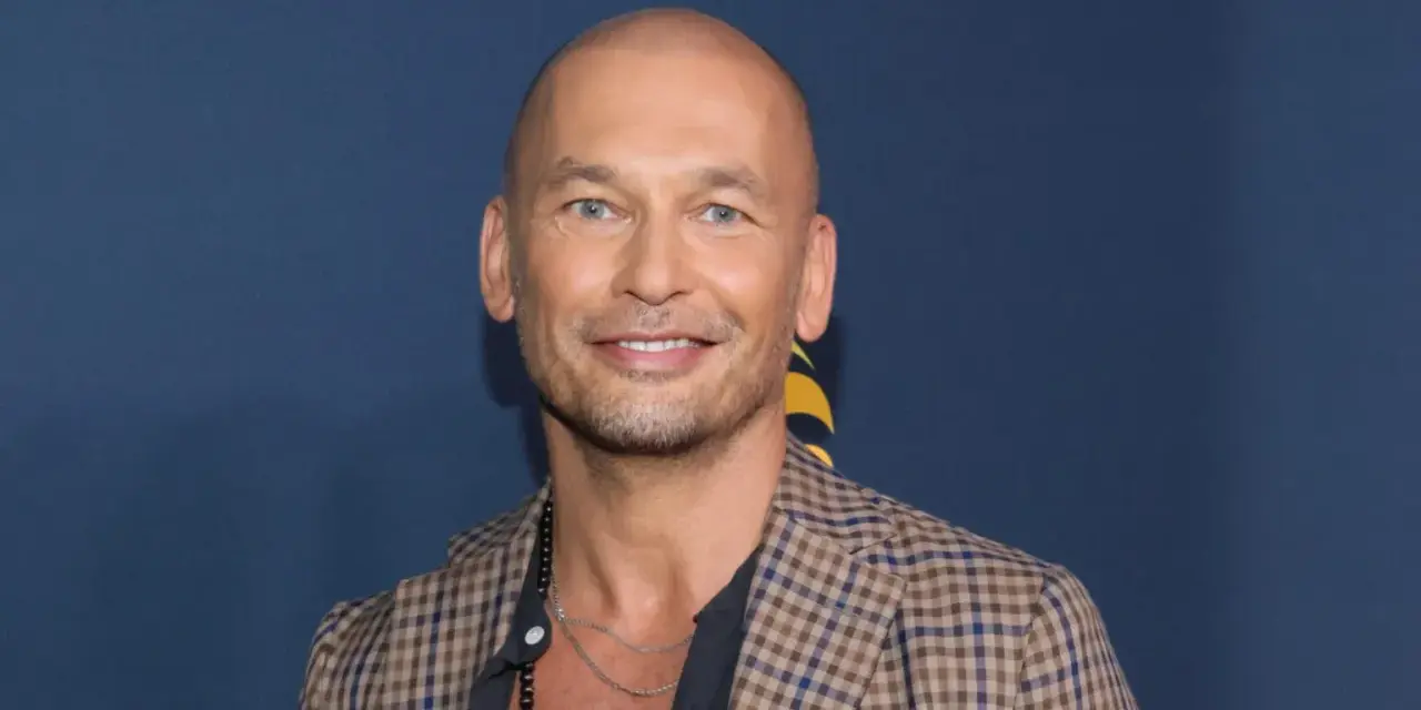 Tomasz Wygoda, łysy mężczyzna z niebieskimi oczami, uśmiecha się, ubrany w kratowaną marynarkę i naszyjnik.