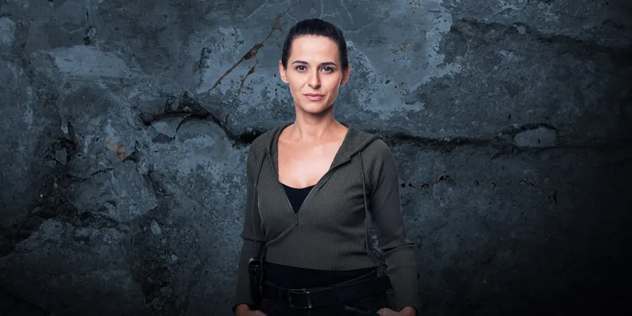 Natalia Brudniak w wojskowym stylu, z rękami w kieszeniach, na tle zniszczonej ściany.