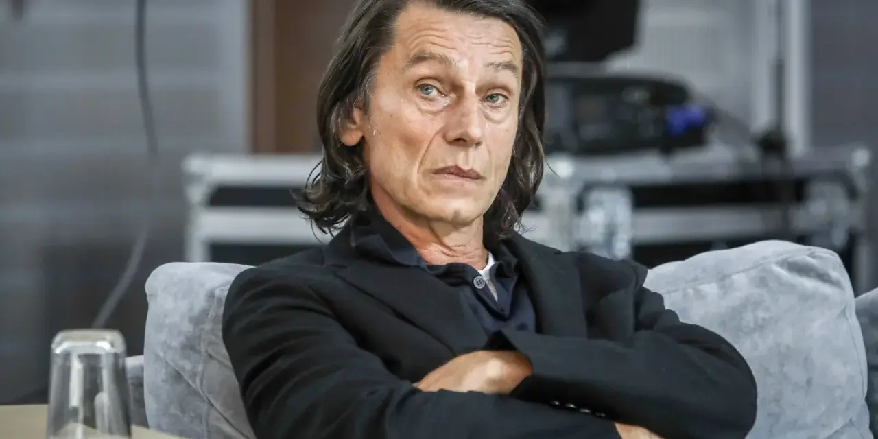 Krzysztof Warlikowski, reżyser teatralny, w czarnym garniturze, siedzi na kanapie. Jego życie prywatne budzi zainteresowanie.
