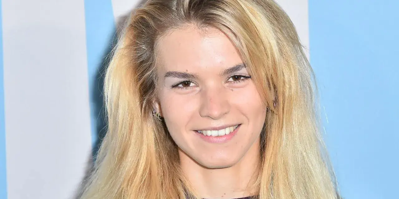 Uśmiechnięta Marta Staśmierska z długimi blond włosami, w fioletowej bluzce, na tle niebiesko-białych pasów.