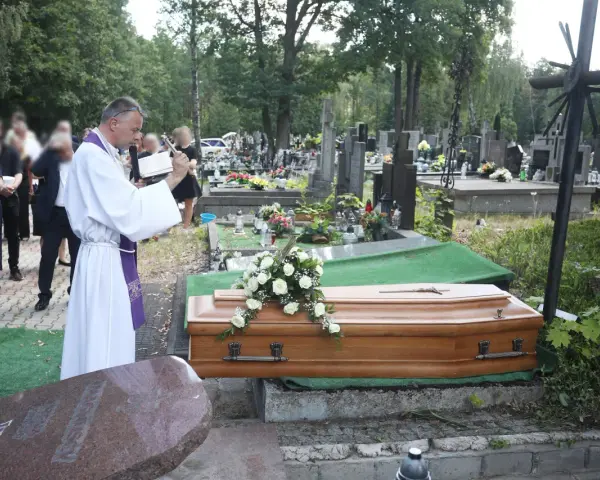 Wojciech Magnuski żegna zmarłego na cmentarzu. Ksiądz w białej szacie czyta z księgi przy trumnie ozdobionej białymi różami.