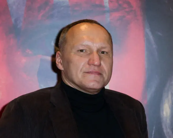 Portret Mariusza Grzegorzka w czarnym golfie i brązowej marynarce na tle abstrakcyjnego wzoru.