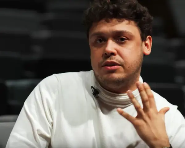 Mężczyzna z kręconymi włosami, Jakub Skrzywanek, w białym golfie gestykuluje podczas rozmowy.