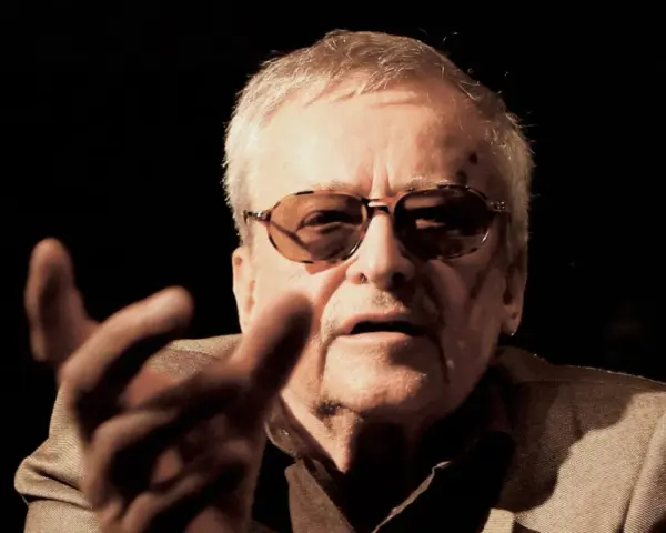 Jerzy Jarocki w okularach, z gestem dłoni, w ciemnym tle.