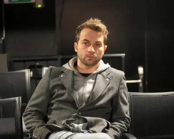 Marcin Hycnar w szarej marynarce i bluzie z kapturem siedzi na czarnym krześle, wpatrując się wprost przed siebie.