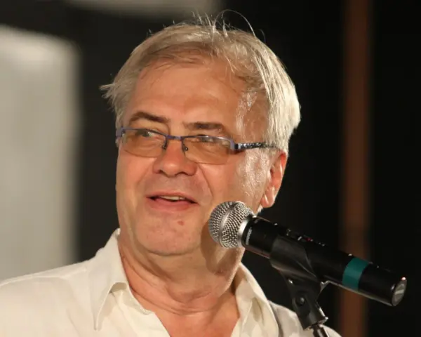 Andrzej Błaszczak, aktor, w okularach i białej koszuli, przemawia do mikrofonu.