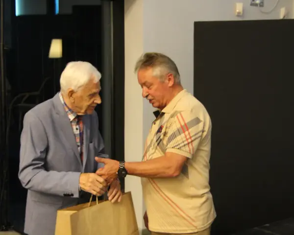 Zdzisław Kuźniar syn, starszy pan w niebieskiej marynarce, wręcza prezent młodszemu mężczyźnie w beżowej koszulce.