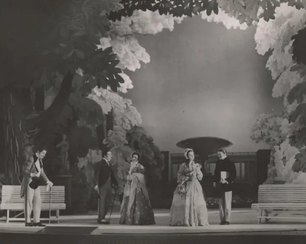 Scena z przedstawienia teatralnego, gdzie aktorzy w strojach z epoki odgrywają scenę w ogrodzie. Reżyser teatralny Szyfman zadbał o szczegóły scenografii.