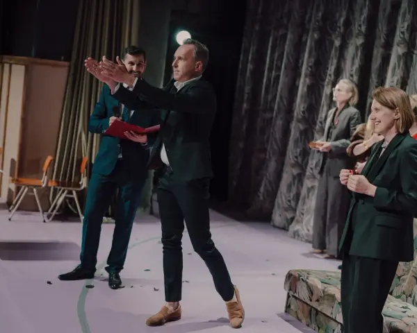Arkadiusz Brykalski w garniturze gestykuluje na scenie, obok niego mężczyzna z teczką. W tle kobiety w eleganckich strojach.
