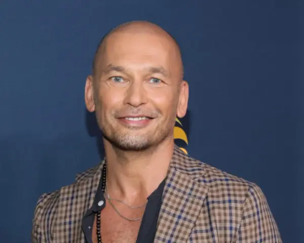 Tomasz Wygoda, łysy mężczyzna z niebieskimi oczami, uśmiecha się, ubrany w kratowaną marynarkę i naszyjnik.