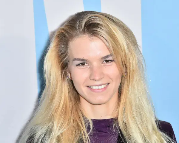Uśmiechnięta Marta Staśmierska z długimi blond włosami, w fioletowej bluzce, na tle niebiesko-białych pasów.