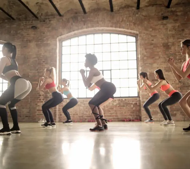 Grupa kobiet ćwiczy w rytm muzyki, wykonując przysiady. To właśnie zumba – świetna zabawa i trening w jednym!