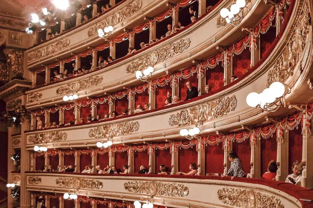 Widok na luksusowe loże teatru La Scala, wypełnione widzami. Czerwone zasłony i złote zdobienia tworzą atmosferę elegancji.