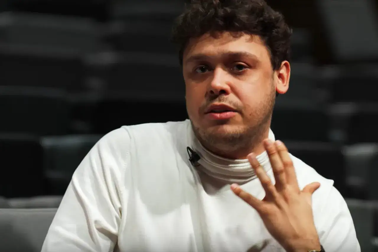 Mężczyzna z kręconymi włosami, Jakub Skrzywanek, w białym golfie gestykuluje podczas rozmowy.
