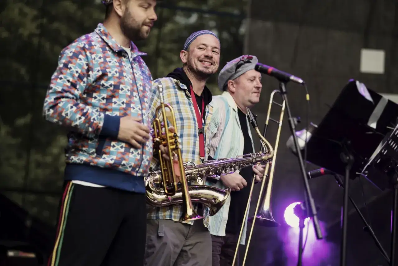 Adam Kłosiński z zespołem na scenie. Muzycy grają na saksofonie, trąbce i puzonie.