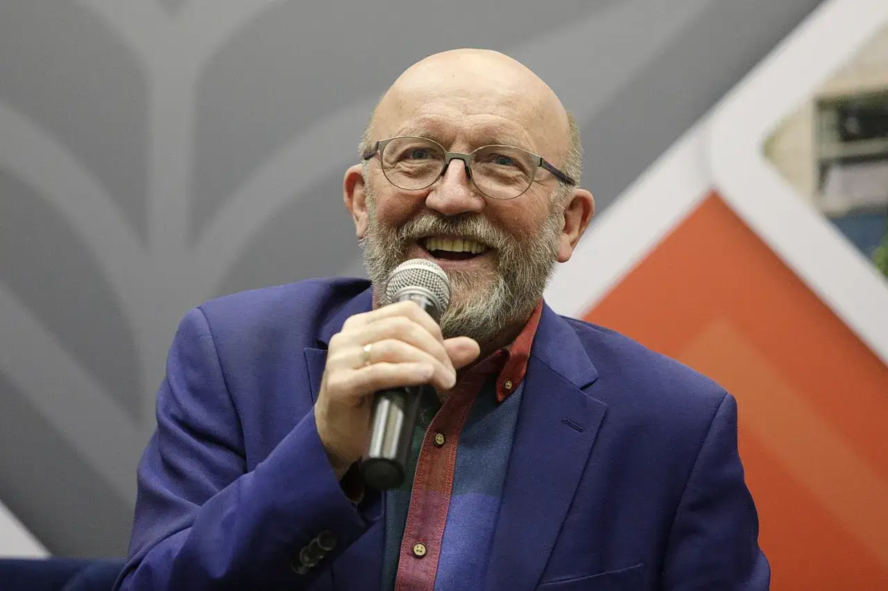 Artur Barciś z uśmiechem trzyma mikrofon, ubrany w niebieską marynarkę i kolorową koszulę.