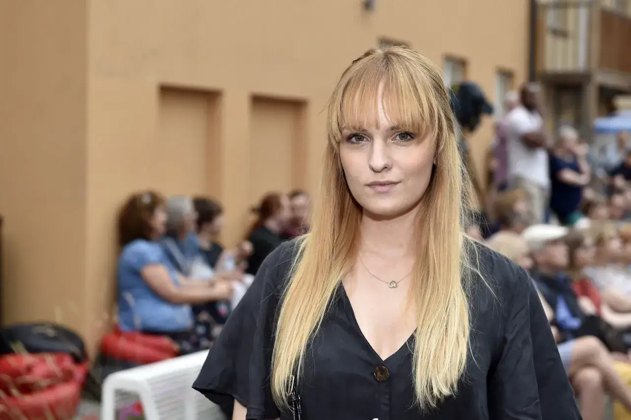 Joanna Biedrzyńska w czarnej bluzce z dekoltem, z blond włosami i grzywką, na tle ludzi.