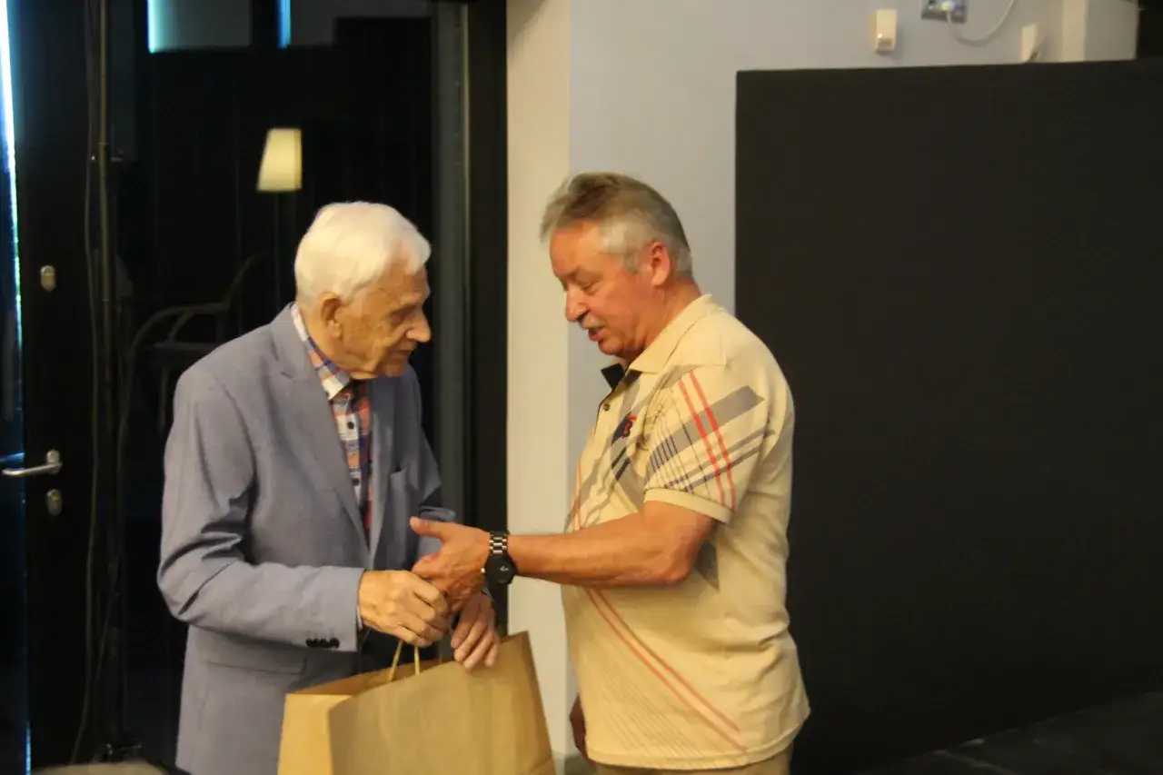 Zdzisław Kuźniar syn, w niebieskiej marynarce, odbiera prezent od starszego mężczyzny w beżowej koszulce.