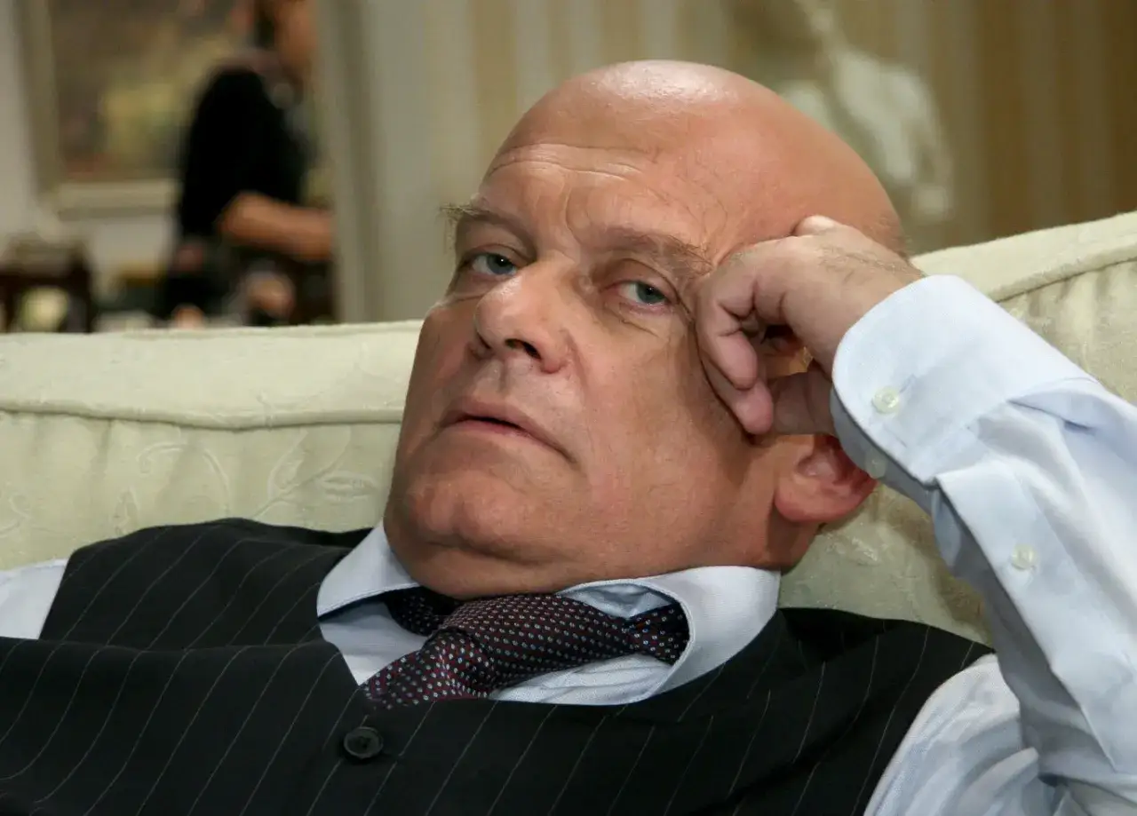 Łysy mężczyzna, znany aktor, opiera głowę na dłoni, ubrany w kamizelkę i krawat.