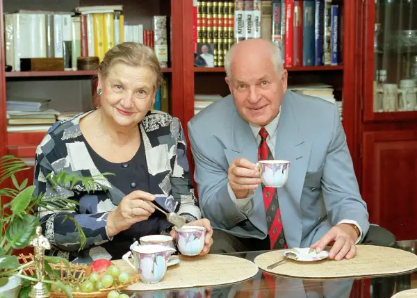 Starsza kobieta i mężczyzna w garniturze piją kawę przy stole, na którym leżą owoce i książki.